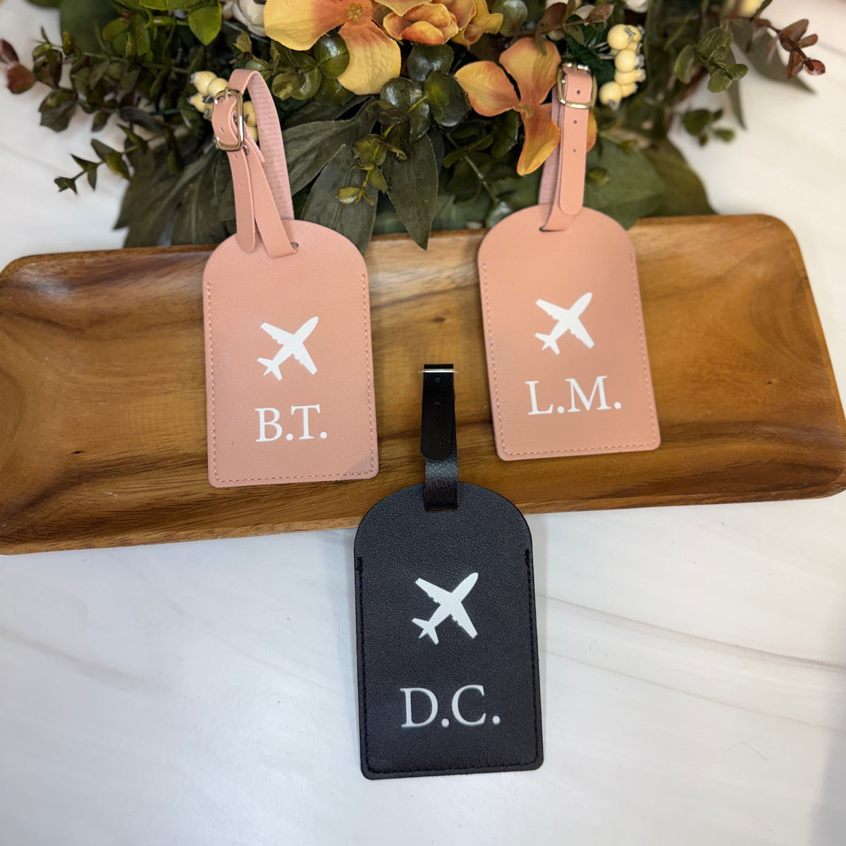 Three luggage tags with initials on a wooden board with flowers in the background