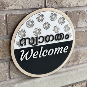 Round bilingual Malayalam and English welcome sign featuring raised 3D lettering, intricate floral mandala patterns, natural wood border, and black-and-white design.