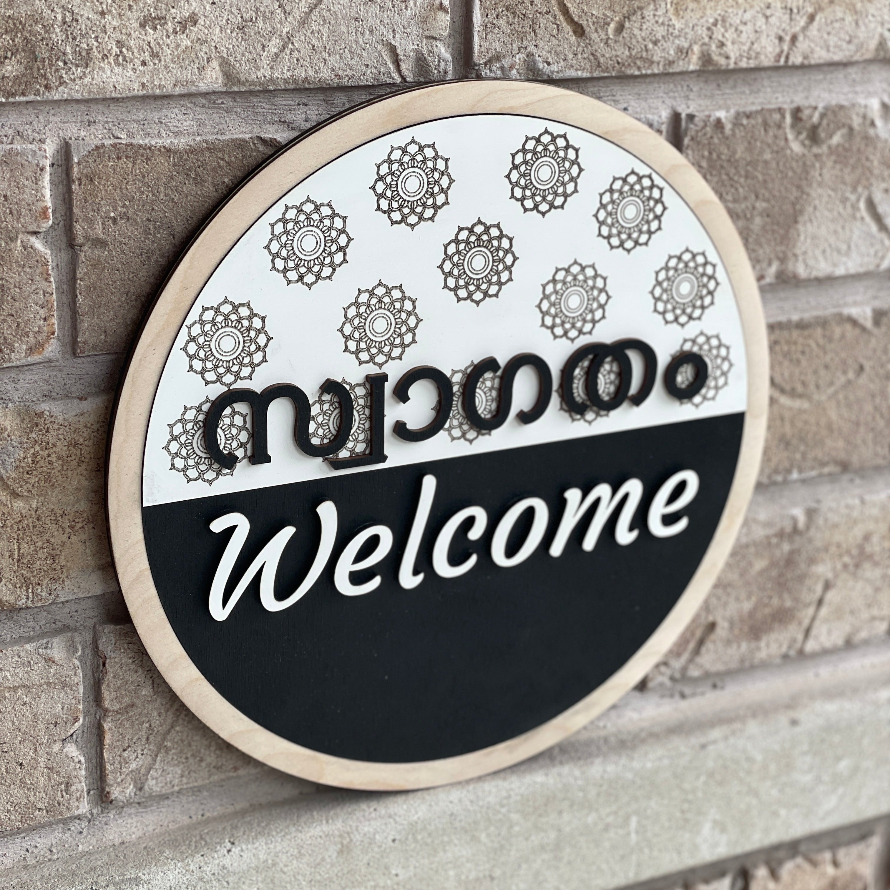 Round bilingual Malayalam and English welcome sign featuring raised 3D lettering, intricate floral mandala patterns, natural wood border, and black-and-white design.