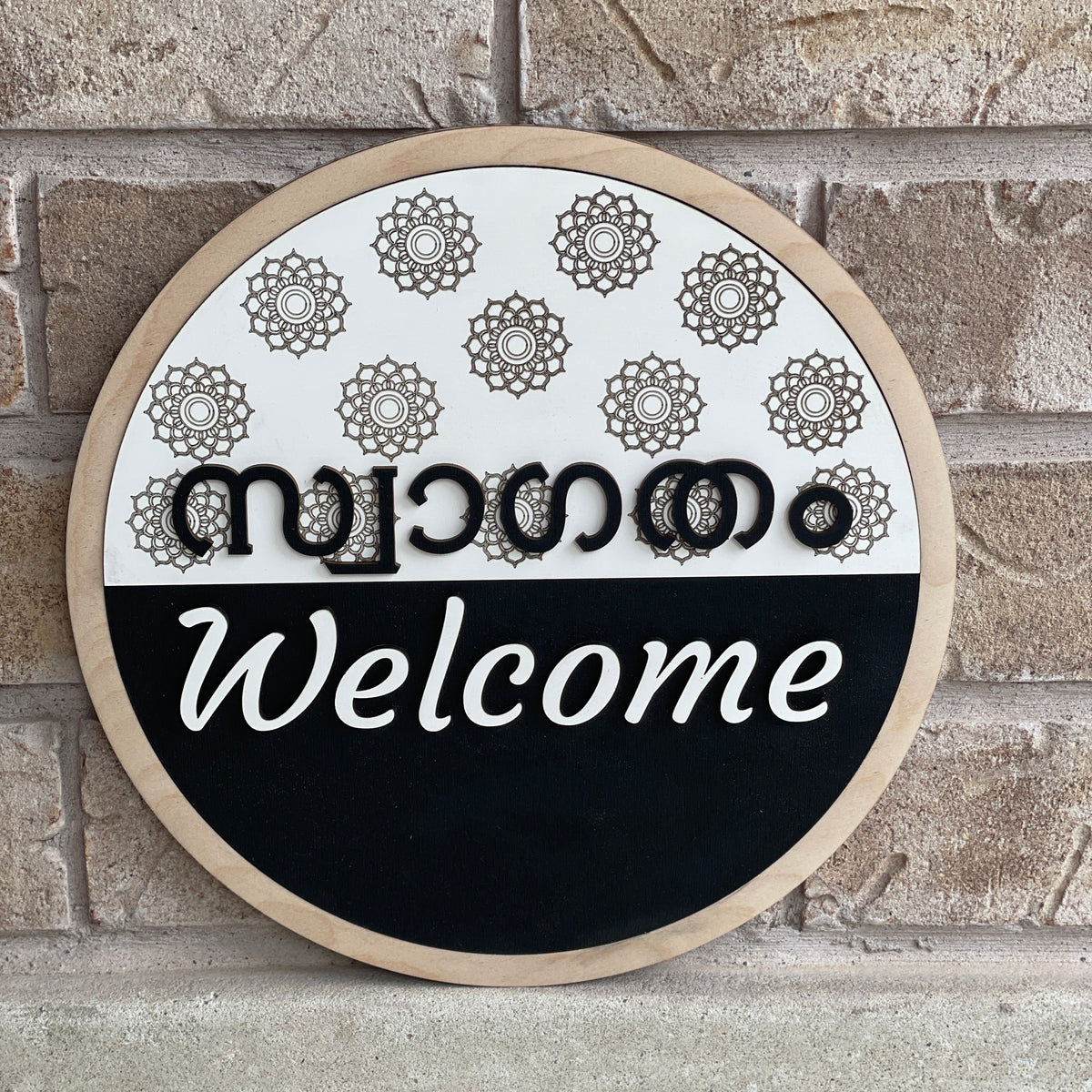Round bilingual Malayalam and English welcome sign featuring raised 3D lettering, intricate floral mandala patterns, natural wood border, and black-and-white design.