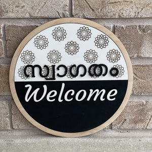 Round bilingual Malayalam and English welcome sign featuring raised 3D lettering, intricate floral mandala patterns, natural wood border, and black-and-white design.