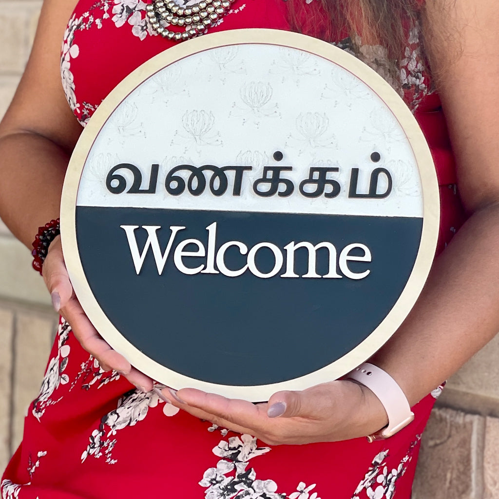 Round Tamil and English bilingual Vanakkam welcome sign with raised 3D lettering, natural wood border, floral background, and black-and-white finish, handcrafted by Timeless Notions.
