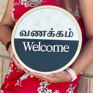 Round Tamil and English bilingual Vanakkam welcome sign with raised 3D lettering, natural wood border, floral background, and black-and-white finish, handcrafted by Timeless Notions.